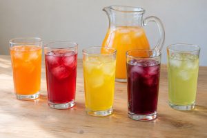 Five tall glasses of iced drinks in orange, red, yellow, dark purple and pale green, with a clear glass jug of orange drink behind, on a light wooden table.